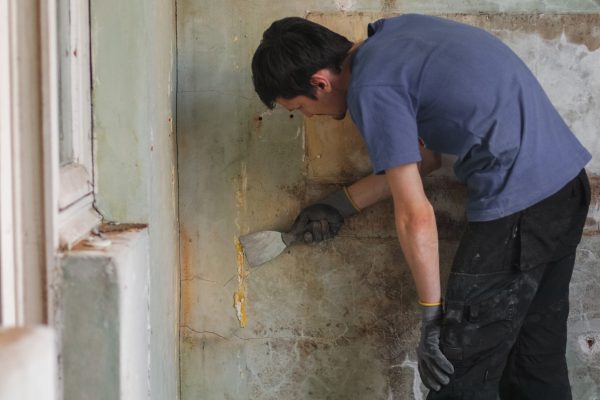One young Caucasian brunette man in uniform cleans the wall with one gloved hand using a spatula in an old and dirty house preparing for renovation, side view close-up.